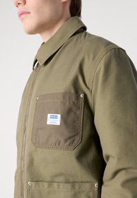 Olive green work jacket with a pointed collar, zipper closure, and a pocket featuring a blue and white label. Durable fabric with visible stitching.