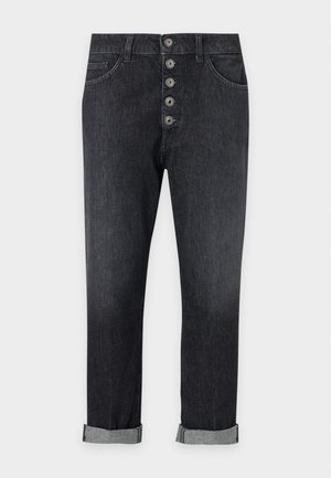 Frau steht mit den Händen in den Taschen, trägt ein bordeauxrotes Kurzarmshirt, schwarze Jeans mit Knopfleiste und Umschlägen sowie weiße Sneakers vor einem schlichten Hintergrund.