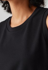 Gros plan sur une personne portant un haut noir sans manches, montrant le cou, l'épaule et les détails du tissu texturé.