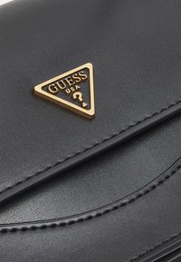 Musta synteettinen käsilaukku, jossa on kolmiomainen kultainen logo, jossa lukee "GUESS USA", sekä sileä tekstuuripinta ja ommeltu yksityiskohta.