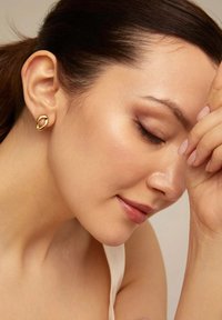 Pendientes de botón de oro con un diseño ovalado, que presentan una textura lisa y un acabado pulido, usados por una modelo con el cabello recogido.