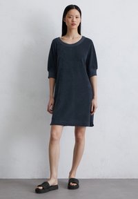 Donkerblauwe katoenen jurk met een ronde halslijn en mouwen tot op de elleboog, met een zachte textuur en een ontspannen silhouet. Zwarte instapsandalen.