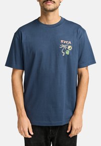 Camiseta de algodón azul con el logo "RVCA" en rosa, y elementos de diseño gráfico en verde y gris en el área del pecho izquierdo. Mangas cortas.