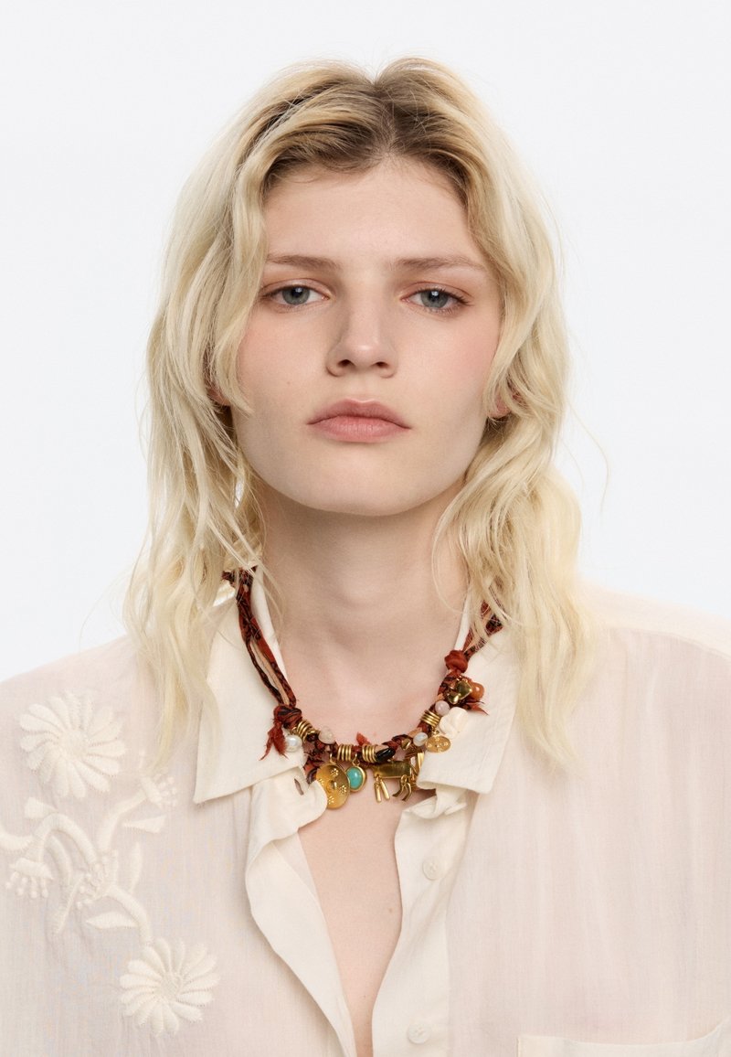 Collar multicolor con cuentas, monedas y acentos sobre un cordón trenzado marrón; se lleva sobre una camisa color crema bordada.