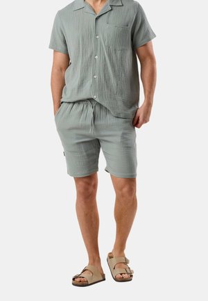 Man draagt een lichtgroen, gestructureerd overhemd met korte mouwen en knopen, en bijpassende short met trekkoord, met beige sandalen met dubbele band.