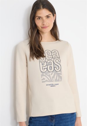 Kvinde med langt brunt hår iført en cremefarvet langærmet skjorte med marineblåt tekst og blå jeans, stående mod en hvid baggrund.