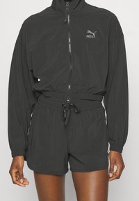 Chaqueta deportiva negra con cuello alto, cremallera y cintura elástica. Combinada con pantalones cortos a juego, que presentan un detalle de logo en el pecho.