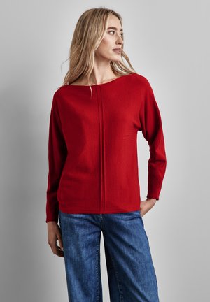 Rode trui met een brede halslijn, lange mouwen en een centrale naaddetail, gecombineerd met relaxte blauwe jeans. Eenvoudige, zachte textuur.
