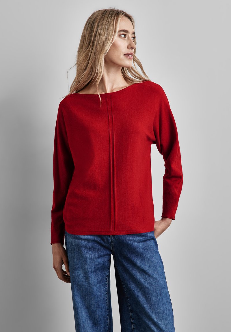 Röd tröja med vid halsringning, långa ärmar och en central sömdetalj, kombinerad med avslappnade blå jeans. Enkel, mjuk textur.