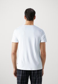 T-shirt en coton blanc à manches courtes et encolure ronde, avec un petit logo à l'ourlet. Le mannequin porte un pantalon de pyjama à carreaux.