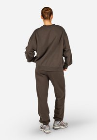 Cilvēks ar matiņu sakrautā bunā stāv pretējā virzienā, uzvilcis brīvu tumši brūnu sporta krekls un bikses, kā arī ar baltām un sudraba sporta apaviem.