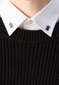 Schwarzer, gerippter Strickpullover mit einem weißen Kragenhemd. Verfügt über metallische Logoakzente am Kragen und an den Knöpfen. Sichtbares Texturdetail im Stoff.