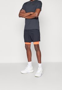Hombre de pie con los brazos cruzados, vestido con una camiseta gris oscura, pantalones cortos negros con ribete naranja, calcetines blancos y zapatillas deportivas blancas.