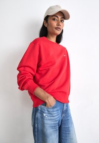 Red sweatshirt with ribbed cuffs and waistband, featuring a small logo. Paired with light blue distressed jeans and a beige cap.