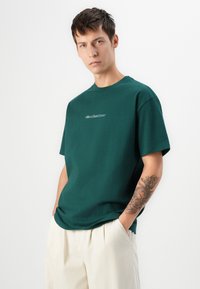 Mann med kort hår iført en grønn Quiksilver t-skjorte og lyse beige bukser, med hendene i lommene, som viser et detaljert tatovering på venstre underarm.