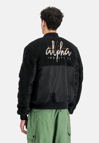 Muž v černé fleecové a nylonové bomber bundě s výšivkou "Alpha Industries" na zádech, v kombinaci se zelenými cargo kalhotami, stojící zády.