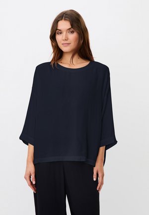 Junge Frau mit langen braunen Haaren, die ein locker sitzendes dunkelblaues Oberteil mit dreiviertel Ärmeln und schwarze Hosen trägt, steht vor weißem Hintergrund.