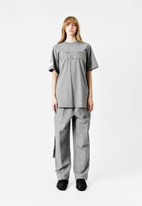 Camiseta oversized gris con logo blanco, combinada con pantalones grises que tienen cremalleras laterales y un corte relajado. Unos zapatos negros texturizados completan el conjunto.