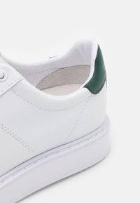 Zapatilla de cuero blanca con talón verde, costuras visibles y suela de goma blanca, mostrada desde la parte trasera sobre un fondo blanco liso.