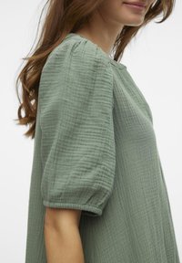 Svijetlozelena haljina teksture s napuhanim rukavima, koja ima opušteni siluetu i okrugli izrez. Tkanina ima mekan, naboran izgled.