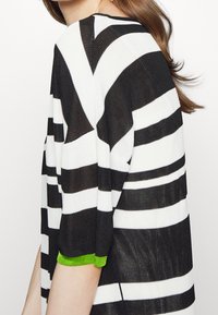 Schwarz-weißer, horizontal gestreifter Pullover mit kurzen Ärmeln und einem grünen Akzent an den Bündchen. Hergestellt aus einem strukturierten Strickstoff.