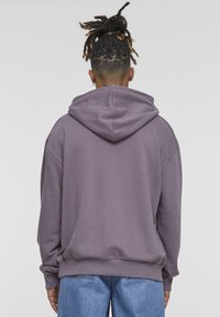 Felpa oversize morbida di colore viola con cappuccio regolabile, maniche lunghe e polsini a costine. Il tessuto appare liscio con una trama uniforme e discreta.