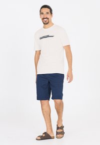 Helle beige T-skjorte med "Yellowstone" grafikk; marineblå shorts med rullekanter; brune sandaler med to stropper og spenner.