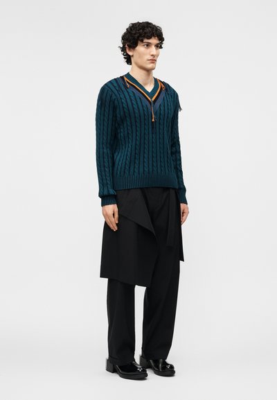 Jeune personne portant un pull à col en V texturé couleur sarcelle, un pantalon foncé et un panneau de jupe noire superposé, assortis à des chaussures à plateforme noires.