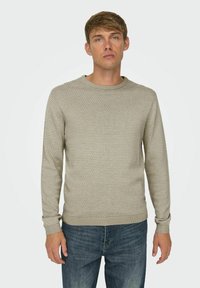 Beige gebreide trui met een textuurpatroon, ronde hals, en geribde manchetten en onderkant. Draagt bij blauwe denim jeans, tentoongesteld op een neutrale achtergrond.