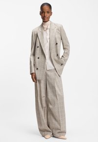 Dobbeltradet beige ternet jakkesætjakke med sorte knapper, kombineret med bukser med vide ben. Under er en lysefarvet skjorte.
