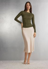 Haut à manches longues vert olive associé à une jupe en satin rose clair lisse et fluide, et des talons ouverts blancs avec des accents de nœud.