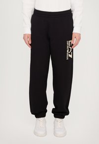 Black sweatpants with an elastic waistband and ribbed cuffs, featuring a cream-colored logo on the left leg. Worn with white sneakers.