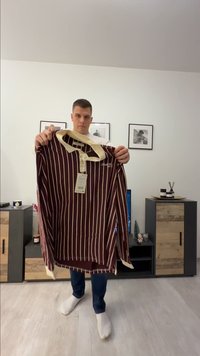 Hombre con calcetines blancos y jeans sosteniendo una camisa polo de manga larga a rayas marrón y beige con una etiqueta en una sala de estar.