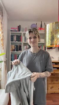 Felpa grigia con zip, indossata da una persona, caratterizzata da una trama morbida e un taglio casual. Sullo sfondo ci sono scaffali di libri e illuminazione decorativa.