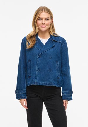 Jonge vrouw die een blauw double-breasted spijkerjasje draagt over een wit shirt en zwarte broek, staand tegen een effen witte achtergrond.