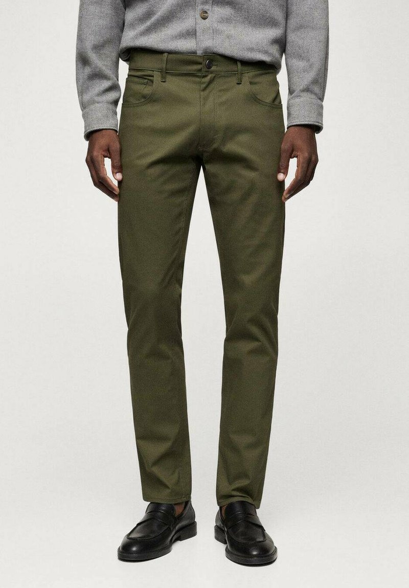 Olivgrüne Chinos mit schlankem Schnitt, Eingrifftaschen und Reißverschluss. Hergestellt aus einem glatten, strapazierfähigen Stoff, kombiniert mit schwarzen Slippern.