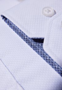 Witte textuuroverhemd met een blauwe gedessineerde binnenvoering en doorzichtige knopen. Close-up van de kraag en de plaatsing van de knopen.