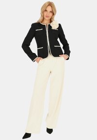 Czarna dopasowana marynarka z białym wykończeniem i kieszeniami; kremowe szerokie spodnie. Duży beżowy kwiatowy akcent zdobi ramię marynarki.