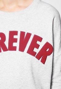 Sudadera gris con llamativo texto en granate que dice "FOREVER" en el pecho. Tejido suave y texturizado con un corte holgado y diseño de cuello redondo.