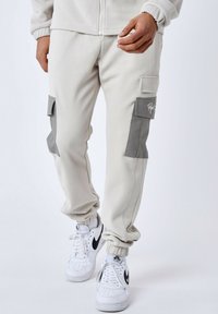 Lichtbeige cargobroeken van gestructureerde stof, met twee grijze klepzakken en elastische boorden, gecombineerd met witte sneakers.