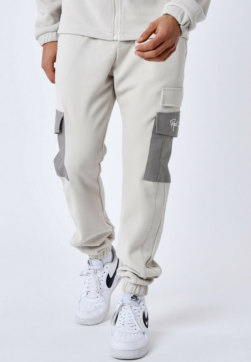 Lichtbeige cargobroeken van gestructureerde stof, met twee grijze klepzakken en elastische boorden, gecombineerd met witte sneakers.