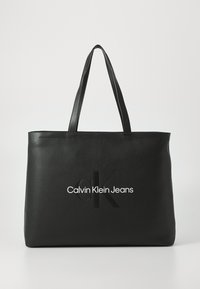 Borsa a spalla nera realizzata in materiale liscio con due manici. Presenta il logo "cK" in rilievo e "Calvin Klein Jeans" in caratteri bianchi sulla parte anteriore.