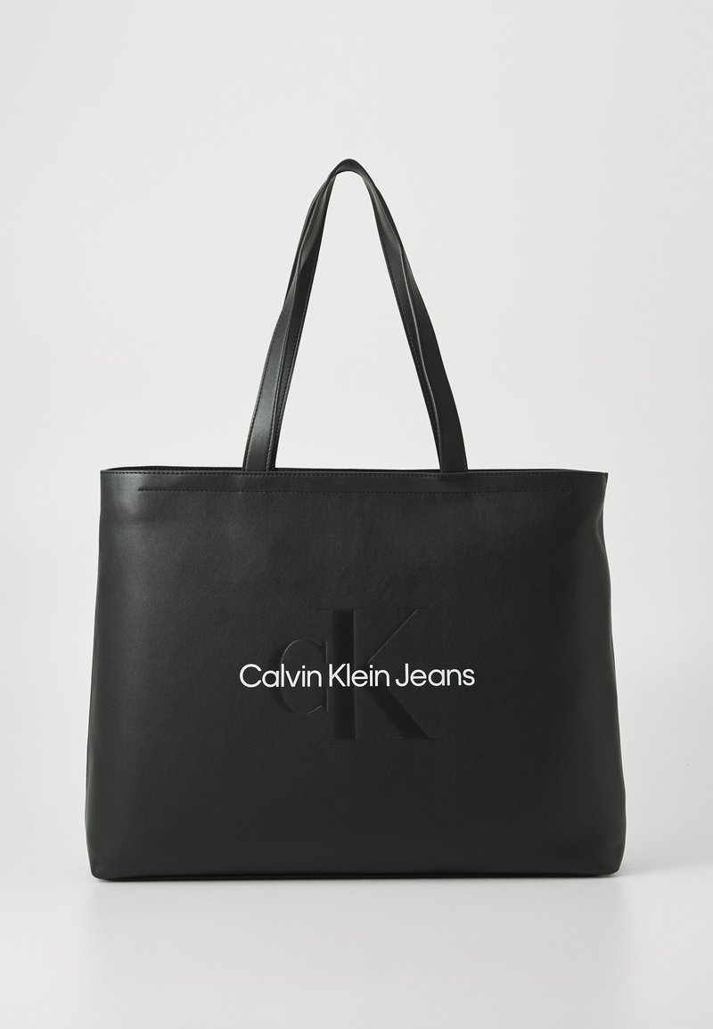 Borsa a spalla nera realizzata in materiale liscio con due manici. Presenta il logo "cK" in rilievo e "Calvin Klein Jeans" in caratteri bianchi sulla parte anteriore.