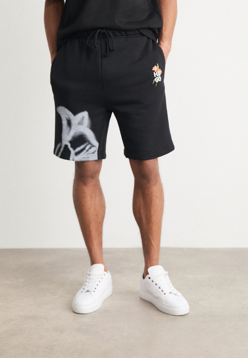Shorts en coton noir avec une coupe décontractée, ornés d'un graphique style graffiti blanc sur le côté gauche et d'un logo floral à l'avant.
