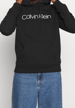 Osoba w czarnej bluzie z kapturem z białym logo "Calvin Klein" i niebieskich dżinsach, ręce regulują bluzę przy szyi.