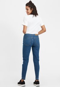 Zilas džinsa bikses ar augstu jostasvietu un taisniem bikšu galiem, ar četriem kabatām un brūnu etiķeti aizmugurē. Balta T-krekls un melni apavi.