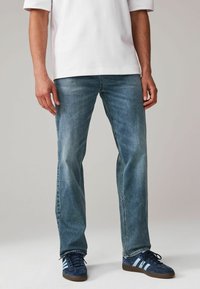 Jeans de mezclilla azul con un corte recto, lavado medio, con un ligero desgaste, combinados con zapatillas de gamuza azul que presentan rayas blancas.