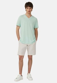 Miętowy t-shirt z dekoltem w serek, miękka tkanina, zestawiony z beżowymi szortami i białymi trampkami. Model stoi swobodnie z rękami w kieszeniach.