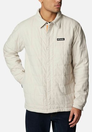 Mann trägt eine hellbeige gesteppte Columbia-Jacke mit Druckknöpfen, steht mit einer Hand in der Tasche und schaut zur Seite.