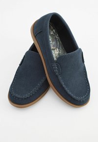 Mørkeblå ruskinds slip-on sko med en tan gummisål, der har dekorativ syning og et mønstret indersål. Glat tekstur og afrundet form.